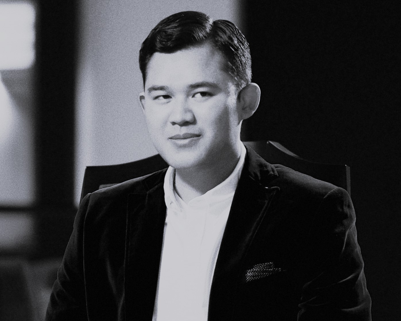 A portrait of a man with short, styled hair, wearing a black blazer over a white shirt, seated in a dimly lit environment.